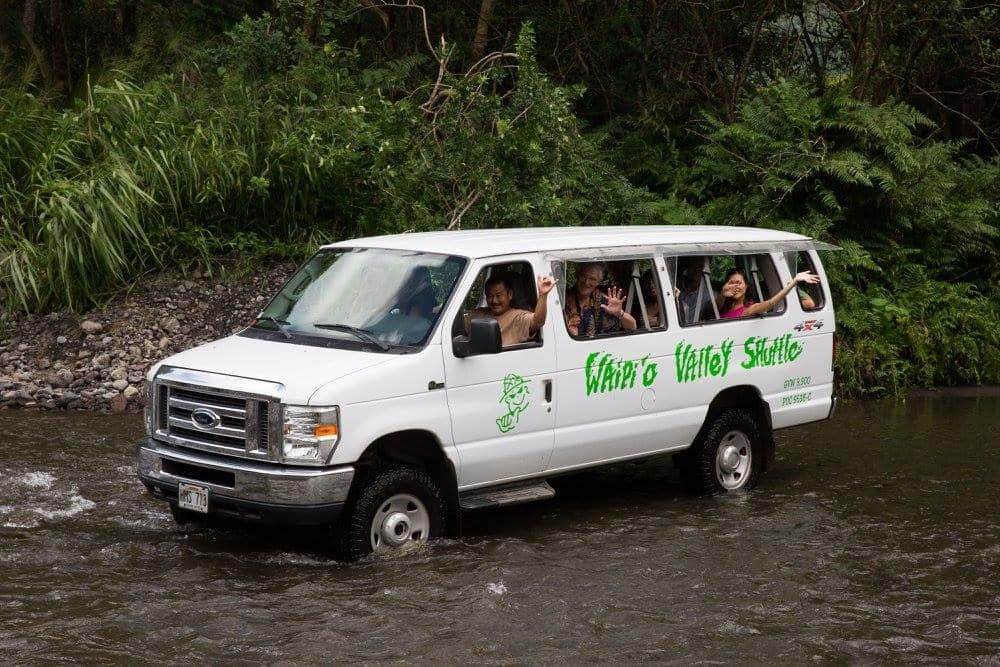 A white van with passengers crossing a shallow river, surrounded by lush green foliage.
