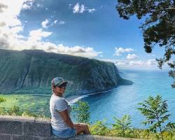 Person sitting on a stone ledge with a view of cliffs and ocean under a blue sky.